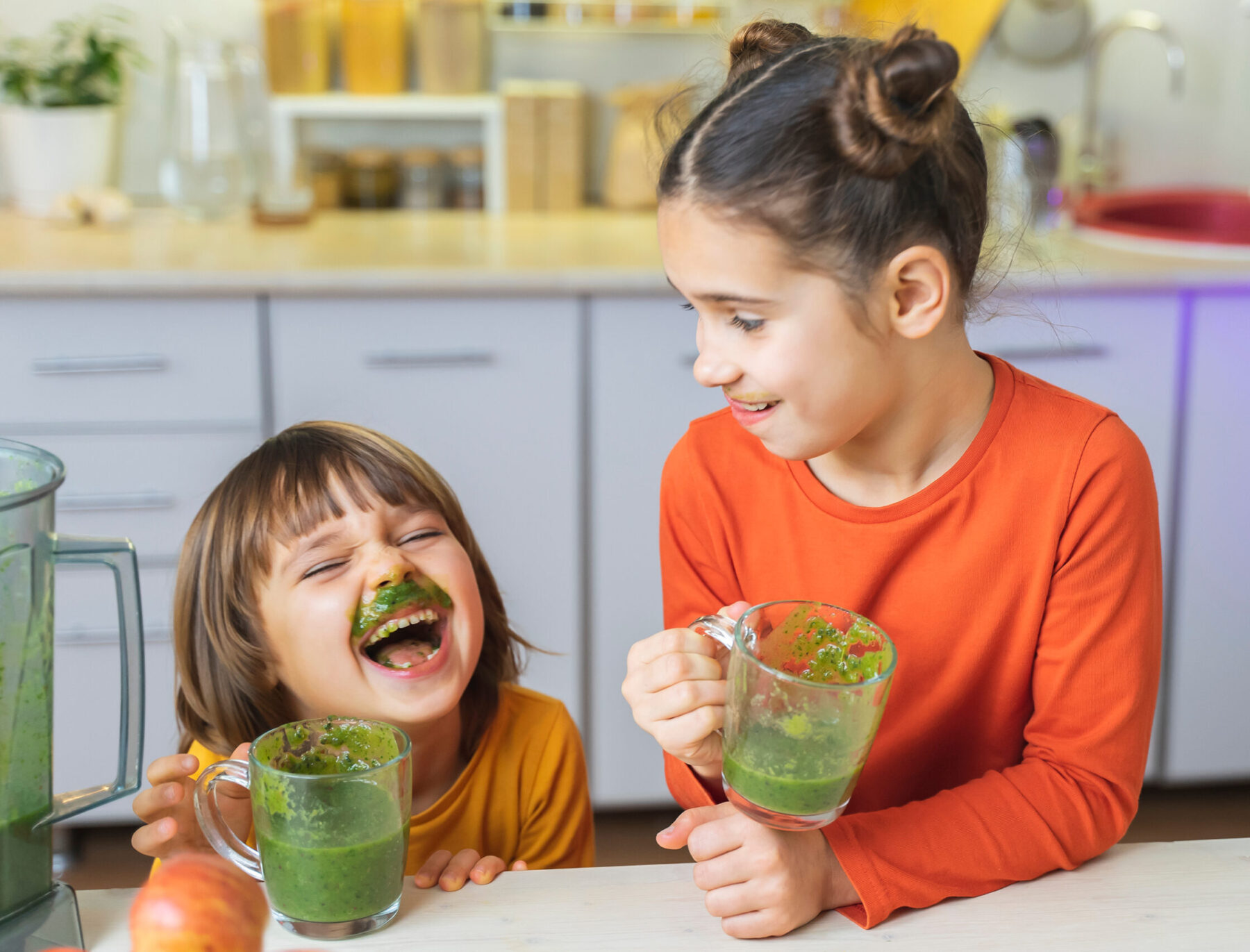 two happy kids wearing orange shirts and drinking a green smoothie in the kitchen