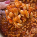 A hand holding a piece of toast with pizza beans on top, above a bowl with the rest of the beans