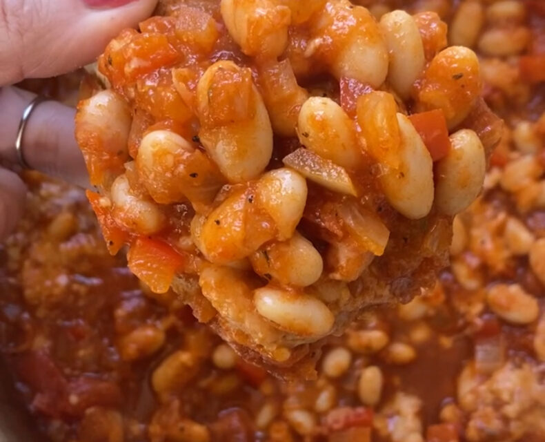 A hand holding a piece of toast with pizza beans on top, above a bowl with the rest of the beans