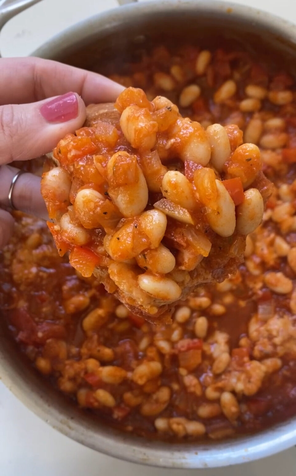 A hand is holding a small piece of bread with pizza beans on top of it, above a pot of the rest of the beans.