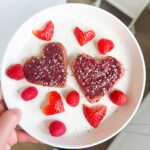 A hand holding a plate of pink heart-shaped pancakes with red strawberry chia jam on top, sprinkled with hemp seeds, surrounded by heart-shaped strawberries and raspberries