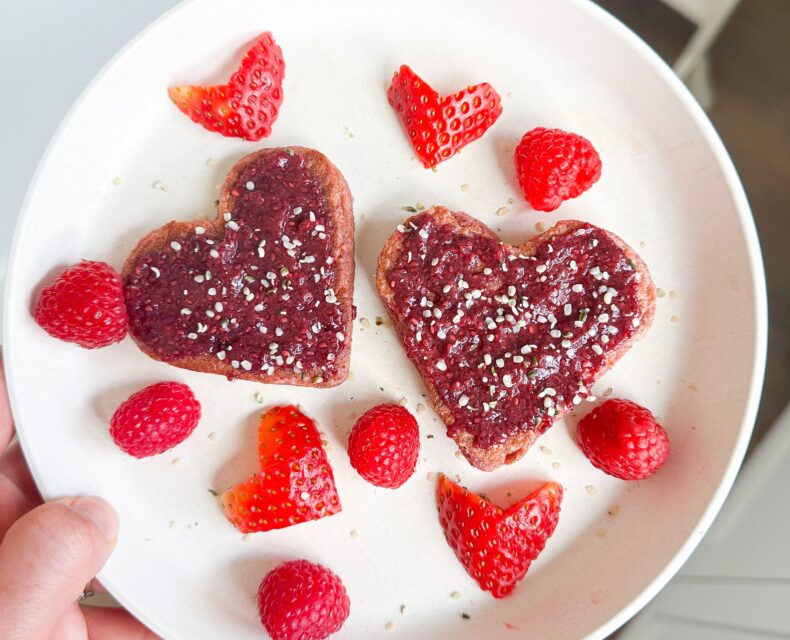 A hand holding a plate of pink heart-shaped pancakes with red strawberry chia jam on top, sprinkled with hemp seeds, surrounded by heart-shaped strawberries and raspberries