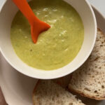 A hand holding plate featuring fresh sourdough bread and a bowl with light green creamy soup with a red spoon in it