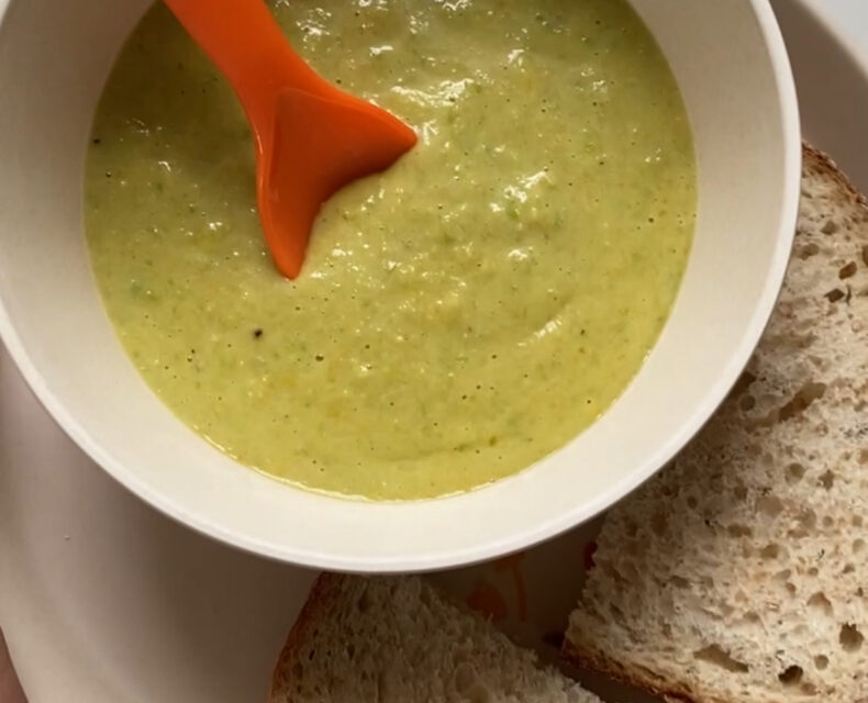 A hand holding plate featuring fresh sourdough bread and a bowl with light green creamy soup with a red spoon in it
