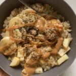A bowl of warm leftover rice mixed with soy milk and cinnamon, and topped with chopped apples, sliced bananas, hemp seeds, chia seeds, and a drizzle of peanut butter and local honey