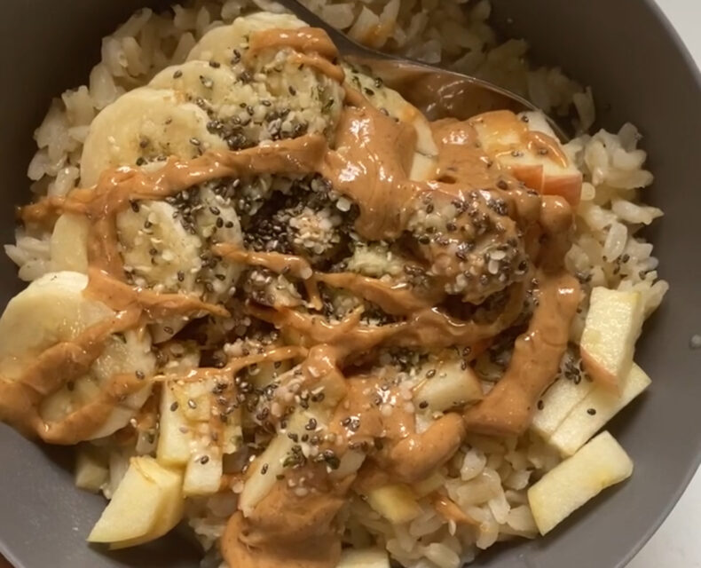 A bowl of warm leftover rice mixed with soy milk and cinnamon, and topped with chopped apples, sliced bananas, hemp seeds, chia seeds, and a drizzle of peanut butter and local honey