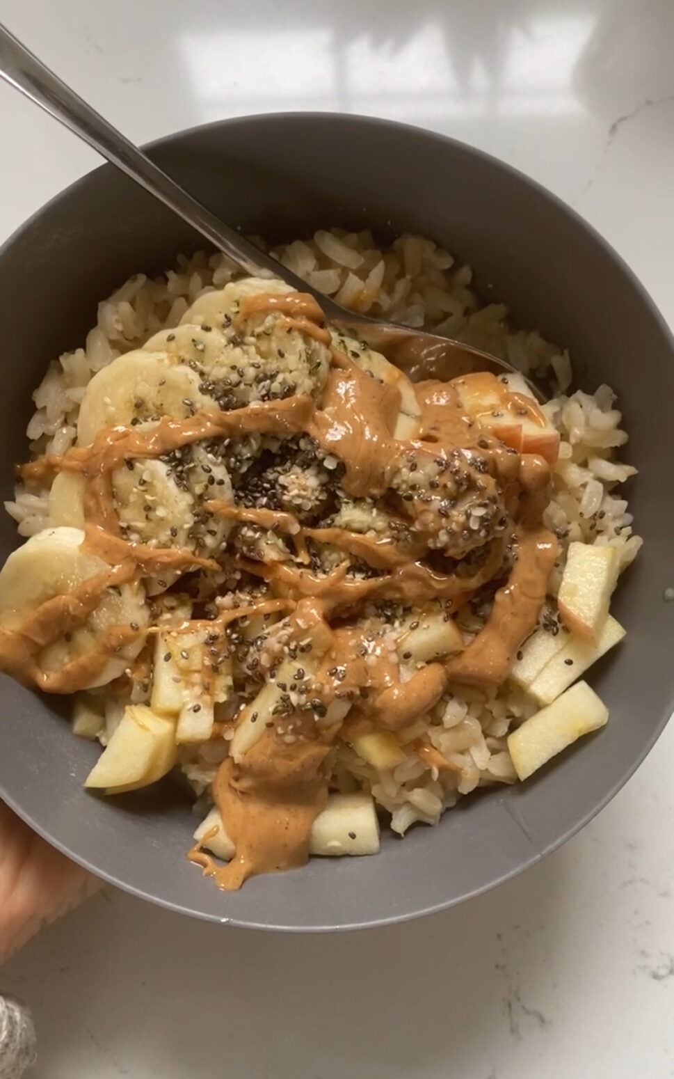 A bowl of warm leftover rice mixed with soy milk and cinnamon, and topped with chopped apples, sliced bananas, hemp seeds, chia seeds, and a drizzle of peanut butter and local honey