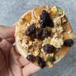 A hand holding a round apple slice topped with peanut butter, raisins, granola, and maple syrup.