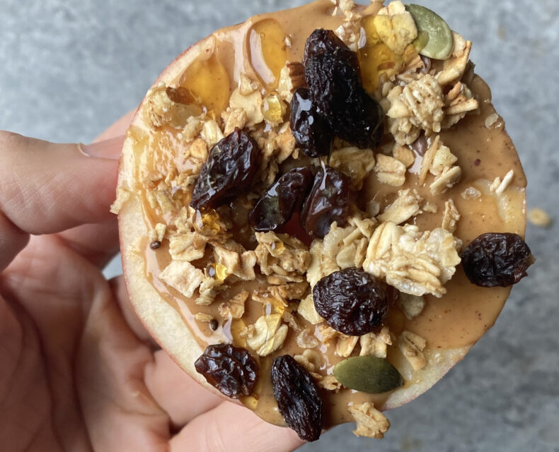 A hand holding a round apple slice topped with peanut butter, raisins, granola, and maple syrup.