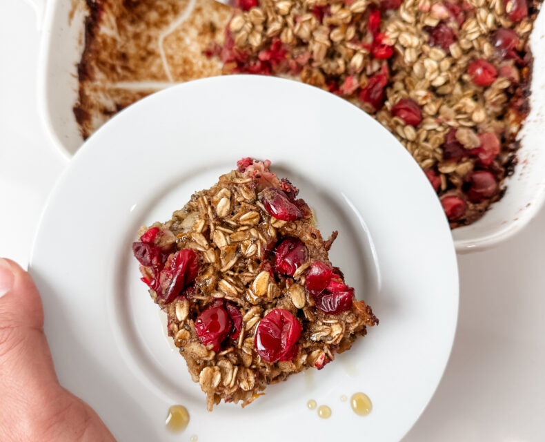 A hand holding a white plate with a square of the Cranberry Orange Oat Bake. Visible on the square are grains of oats and squished baked cranberries. The plate is being held over a casserole dish with the rest of the Cranberry Orange Oat Bake.