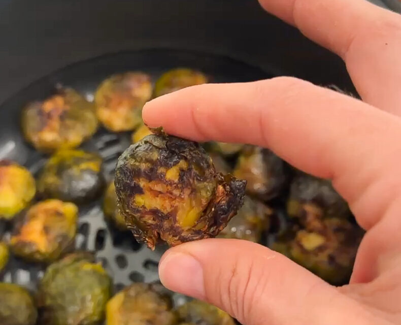 A hand holding a single browned crispy smashed Brussels sprout over an air fryer basket containing the rest of the Brussels sprouts.