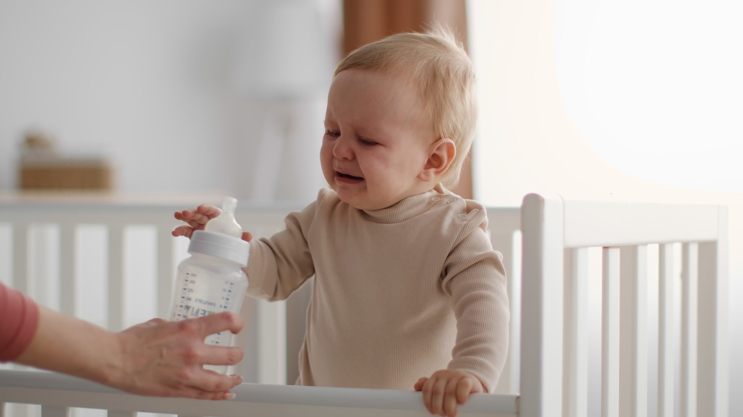 Baby standing up in crib crying and pushing bottle away in mom's hand.