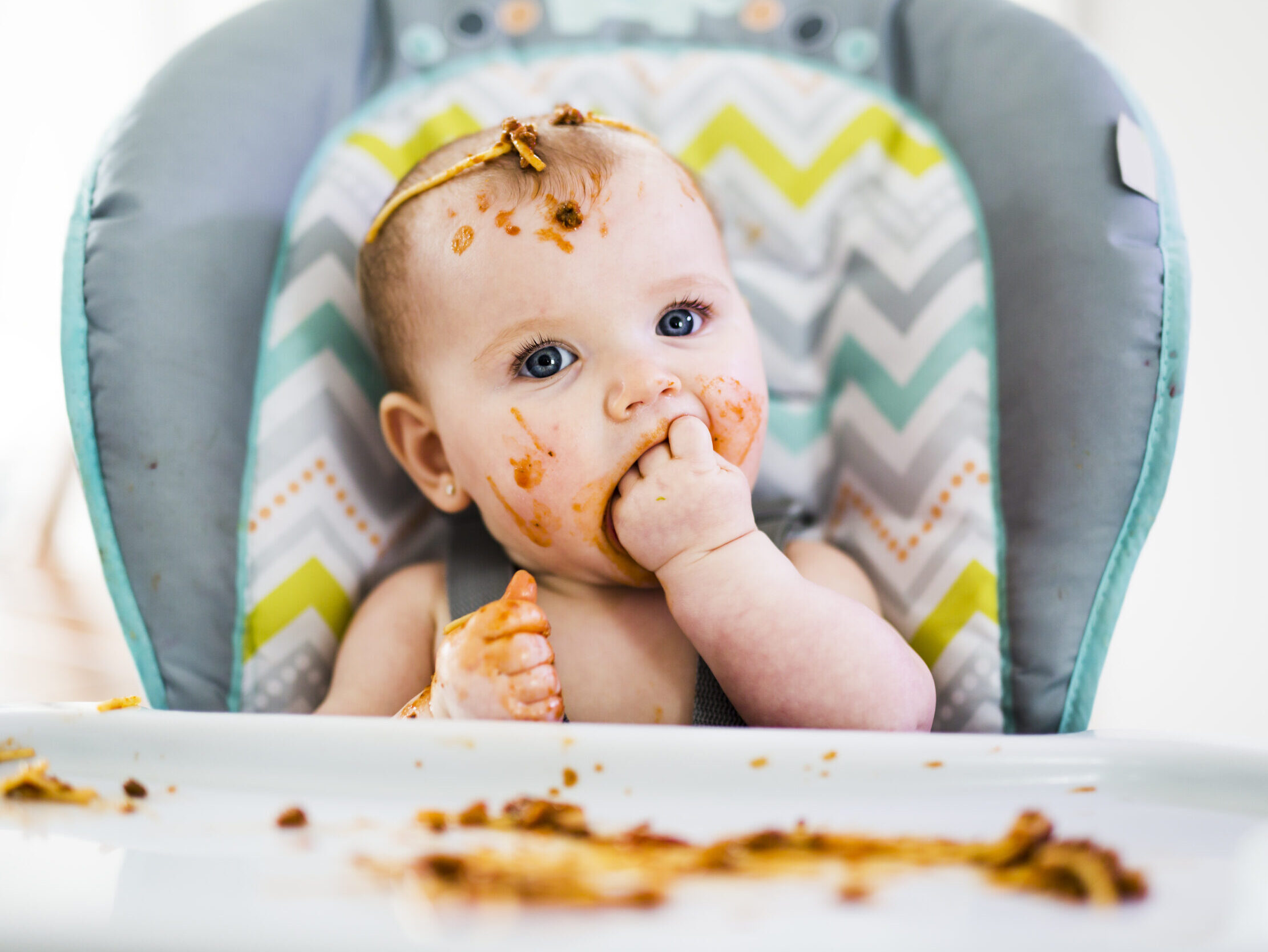 baby sitting in high chair eating best first foods for baby with spaghetti on her head
