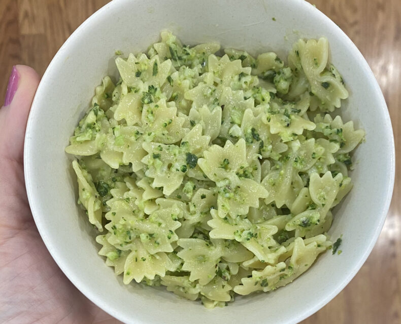A hand holding a white bowl filled with light green bow tie pasta with green flecks on it (from basil and broccoli).