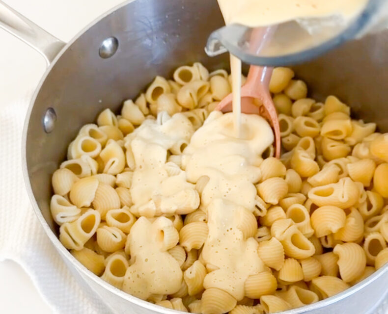 Creamy light yellow sauce is being poured out of a blender into a pot of pasta shells