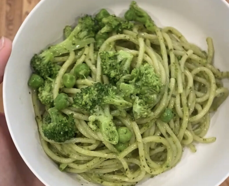 A hand holding a white bowl of green-tinted spaghetti pasta with broccoli and peas mixed in and on top, mixed with pesto sauce.
