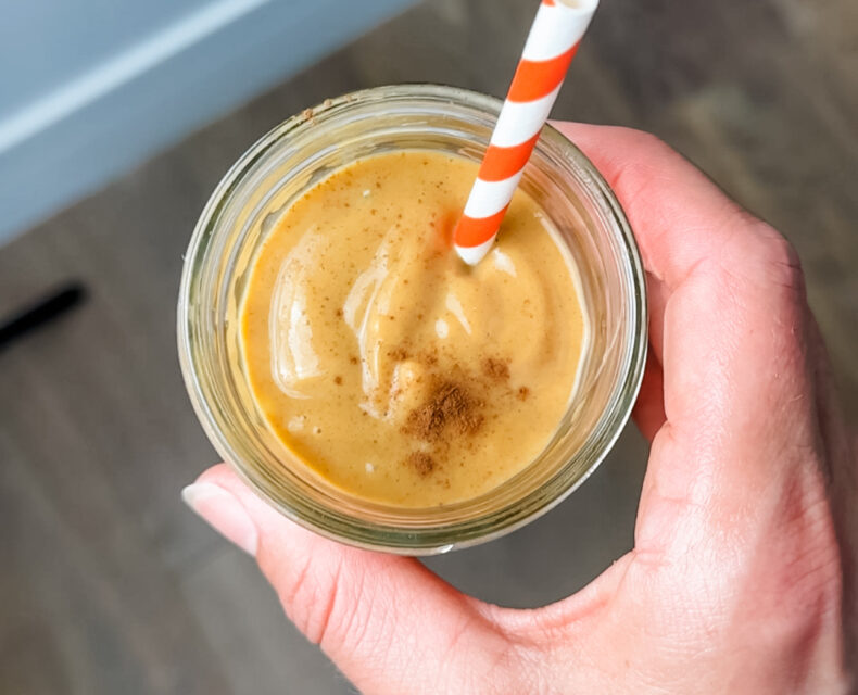 A hand holding a glass of orange sweet potato pie smoothie. There is cinnamon sprinkled on top and a red and white barber pole straw coming out of the glass.
