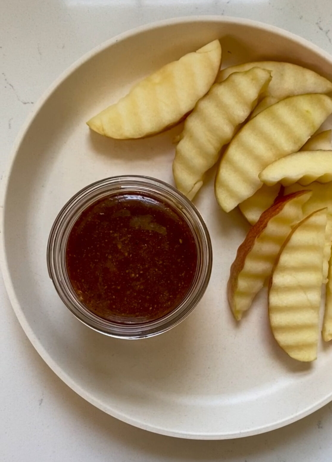 A tan plate with waffle cut apple slices from a red apple, with a round little dip bowl containing caramel sauce.
