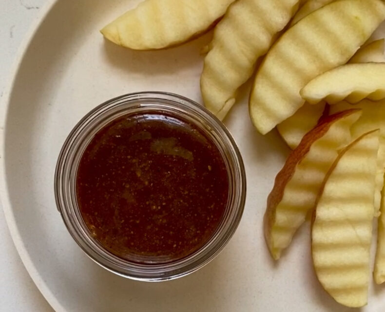 A tan plate with waffle cut apple slices from a red apple, with a round little dip bowl containing caramel sauce.