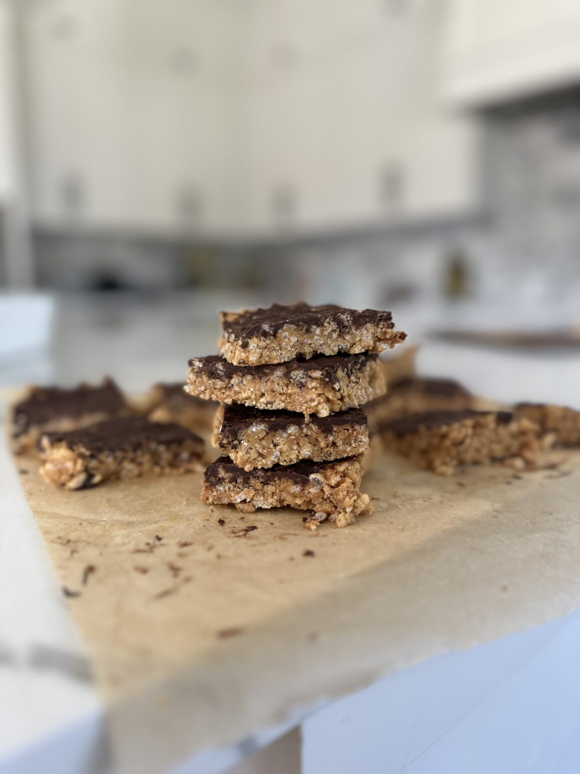a stack of peanut butter chocolate brown rice crispy treats on a table in a white kitchen. Looking at the stack from the side. The treats on sitting on a piece of parchment paper. They look like squares made out of cereal with a coating of chocolate on top.