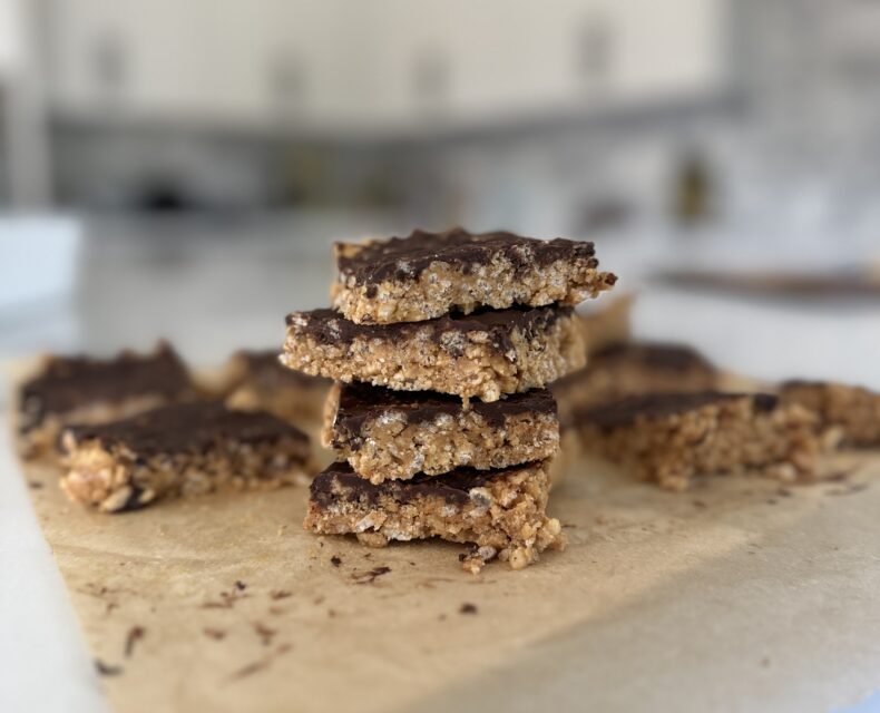 a stack of peanut butter chocolate brown rice crispy treats on a table in a white kitchen. Looking at the stack from the side. The treats on sitting on a piece of parchment paper. They look like squares made out of cereal with a coating of chocolate on top.