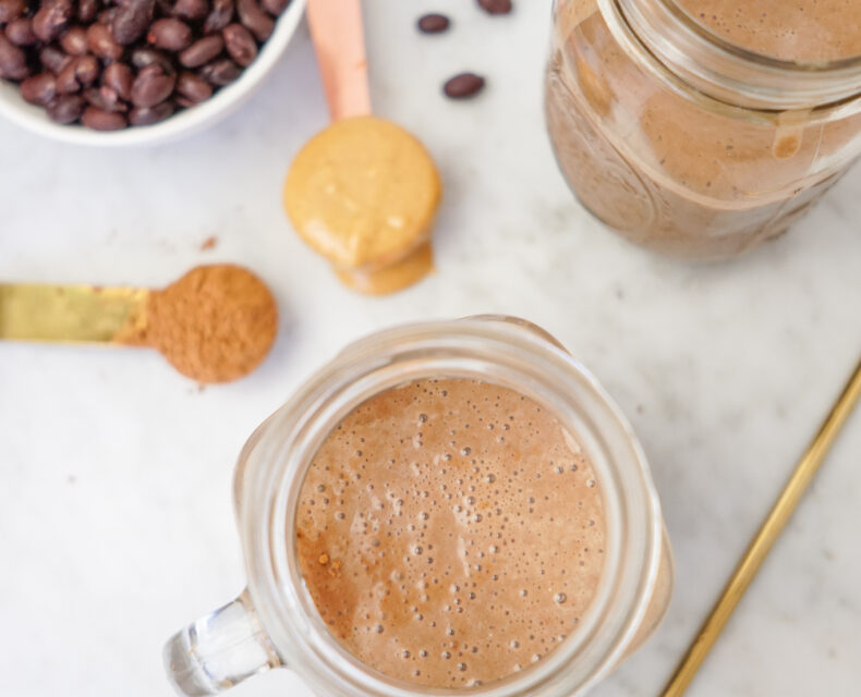 A clear mug containing a foamy light brown chocolately smoothie, with another clean mug of it up and to the right, as well as a bowl of black beans in the upper left, a tablespoon of cocoa powder, and another tablespoon with nut butter. All of this is on a white marble countertop surface.
