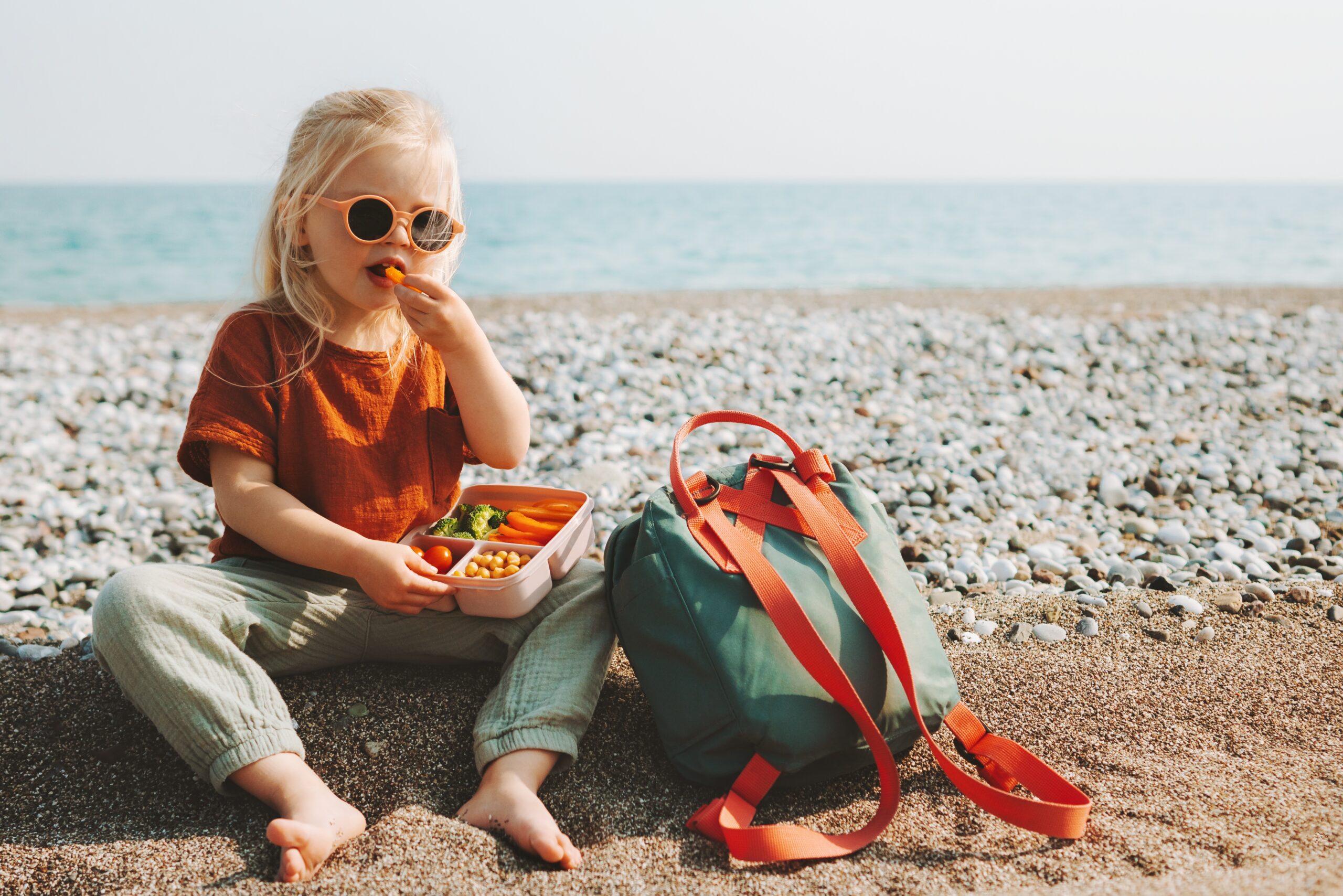 girl sitting on the beach eating veggies out of a tray healthy snacks for summer