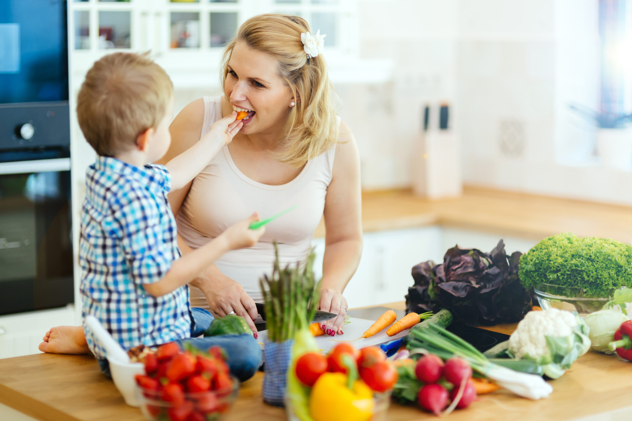 child feeding mother a carrot while sitting on kitchen island surrounded by colorful fruits and vegetables